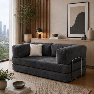Puede incluir: Un sal&oacute;n moderno con un sof&aacute; de pana gris oscuro y un coj&iacute;n blanco. La habitaci&oacute;n incluye una mesa de centro redonda, una alfombra beige y una gran ventana con vistas a la ciudad. Los elementos decorativos incluyen una obra de arte enmarcada y una planta en maceta.