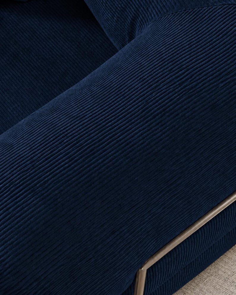 May include: Close-up of a section of a navy blue corduroy sofa. The fabric has a ribbed texture, with visible vertical lines. A silver-colored metal frame is visible on the edge of the sofa.