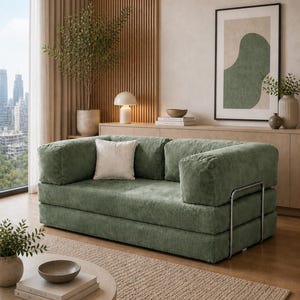 May include: A sage green corduroy sofa with a modern design. The sofa has a low profile, square arms, and a metal frame. A white pillow rests on the sofa. The room features a neutral color palette with a round coffee table and a framed abstract art piece.