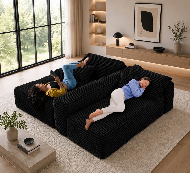 Puede incluir: Sof&aacute; seccional de pana negro de dos piezas con dos personas relaj&aacute;ndose en &eacute;l. Una persona est&aacute; leyendo un libro y la otra est&aacute; durmiendo. El sof&aacute; est&aacute; sobre una alfombra de color claro en un sal&oacute;n moderno.