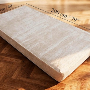 Puede incluir: Colch&oacute;n rectangular beige, de 200 cm de largo, 100 cm de ancho y 18 cm de grosor. El colch&oacute;n tiene una superficie texturizada y est&aacute; colocado sobre un suelo de madera.