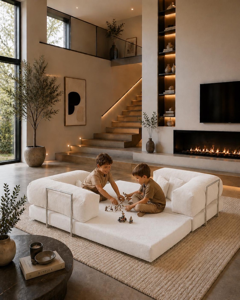 Puede incluir: Dos ni&ntilde;os jugando en un sof&aacute; modular blanco con soportes de metal plateado. El sof&aacute; est&aacute; sobre una alfombra beige en una sala de estar moderna con chimenea, estanter&iacute;as empotradas y una escalera. La habitaci&oacute;n tiene tonos neutros y luz natural.