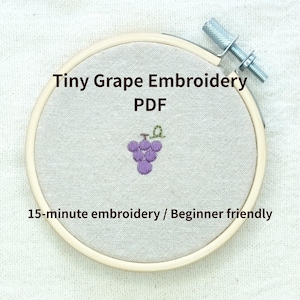 May include: A circular wooden embroidery hoop containing light beige fabric with a small purple grape cluster design. The text Tiny Grape Embroidery PDF and 15-minute embroidery / Beginner friendly is printed over the fabric in dark brown.