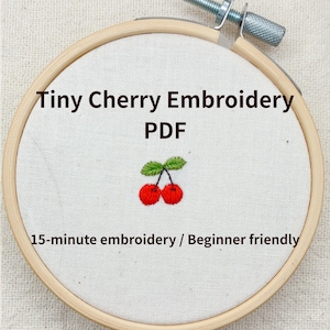 Może przedstawiać: Drewniana tamburyna do haftu trzyma białą tkaninę z małym haftem z wiśniami. Tekst brzmi "Tiny Cherry Embroidery PDF" i "15-minute embroidery / Beginner friendly". Wiśnie są czerwone z zielonymi liśćmi.
