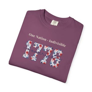 May include: A plum-colored t-shirt featuring the white text One Nation Indivisible above the number 1776. The digits are filled with a red, white, and blue floral pattern, with the word America written in a cursive script across the center.