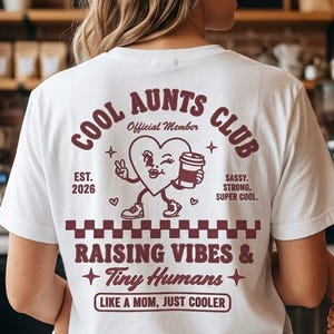 Puede incluir: Una camiseta blanca con un gráfico granate en la espalda con el texto COOL AUNTS CLUB, Official Member, EST. 2026, SASSY. STRONG. SUPER COOL., RAISING VIBES & Tiny Humans, y LIKE A MOM, JUST COOLER, junto a un personaje de corazón.