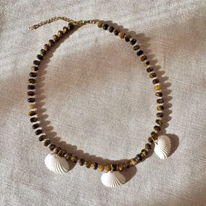 Puede incluir: Un collar de cuentas con piedras de ojo de tigre facetadas en tonos marrón y dorado, intercaladas con pequeñas cuentas transparentes. El collar incluye tres dijes de conchas blancas sujetos con anillas metálicas doradas y una cadena extensora.