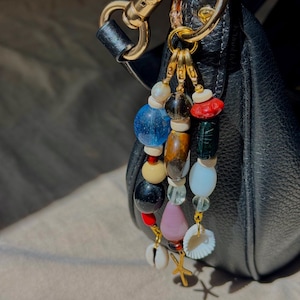 May include: A bag charm featuring three strands of multi-colored beads, including blue, black, brown, pink, and white spheres, accented with small seashells and a gold-toned starfish charm, attached to a gold-toned metal lobster clasp.