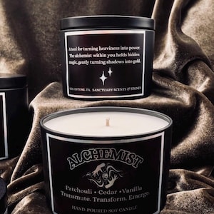 May include: A black soy candle in a metal tin with a white label featuring the text ALCHEMIST, Patchouli, Cedar, Vanilla, Transmute, Transform, Emerge, and NET WT. 8 OZ / 227G. A second black candle tin above displays text about turning shadows into gold.