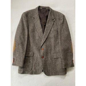 Puede incluir: Una blazer de tweed marrón con coderas color canela y botones marrones. La chaqueta tiene un diseño clásico con dos bolsillos delanteros y solapas con muescas. El forro interior es de color marrón oscuro.