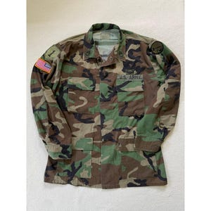May include: Camouflage pattern military jacket with the words "U.S. ARMY" printed on the front. The jacket has multiple pockets, a US flag patch, and other military insignia. The jacket is a mix of green, brown, and black.