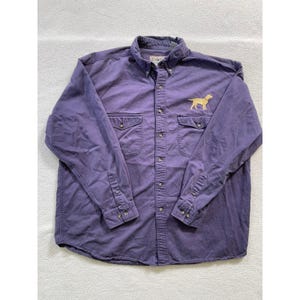 Könnte beinhalten: Ein langärmeliges Button-Down-Hemd in einem gedämpften Violett. Das Hemd verfügt über einen geknöpften Kragen, zwei Brusttaschen mit geknöpften Klappen und eine gestickte goldfarbene Hundesilhouette auf der oberen linken Brustseite.