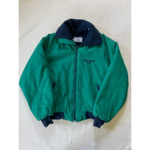 May include: A green jacket with a navy blue collar, cuffs, and trim. The jacket has a full zipper and the words "Performance Coal" embroidered on the front. The jacket is made of a lightweight material.