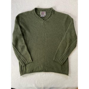May include: An olive green V-neck jumper. The jumper has a ribbed texture and ribbed detailing on the sleeves. A small tag is visible near the neckline. The jumper is made of a soft, knitted material.