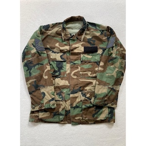 Op de afbeelding: Een camouflagejas met lange mouwen in een bospatroon met tinten bruin, groen en zwart. De jas is voorzien van een knoopsluiting, vier voorzakken met knoopflappen en een zwarte rechthoekige patch op de borst.