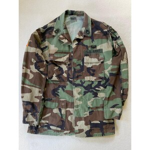 Könnte beinhalten: Militärjacke mit Tarnmuster, Knopfleiste, vier Vordertaschen und Kragen. Auf der Jacke steht der Schriftzug "U.S. ARMY" über einer der Taschen. Die Farbpalette umfasst Grün-, Braun- und Schwarztöne.