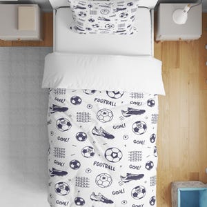 Puede incluir: Juego de cama blanco con un estampado de fútbol en azul marino. El conjunto incluye una funda nórdica y una funda de almohada, ambas con balones de fútbol, zapatillas deportivas y la palabra "GOAL!". Ideal para el dormitorio de un aficionado al deporte.