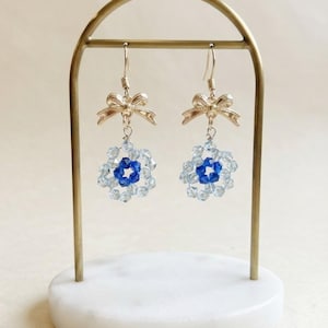 May include: A pair of dangle earrings featuring a gold-toned metal bow at the top, connected to a circular floral-shaped charm made of faceted light blue and dark blue crystal beads, suspended from a gold-toned metal hook earring finding.