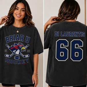 Puede incluir: Camiseta gris oscuro con dise&ntilde;o de hockey, con el texto "Briar U Hockey" y un logotipo de p&aacute;jaro y palos de hockey cruzados. La parte trasera muestra el nombre "Di Laurentis" y el n&uacute;mero "66" en azul marino.