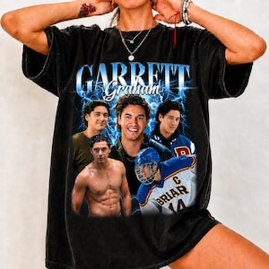 Peut inclure: Un t-shirt noir à manches courtes avec un imprimé graphique portant le texte "GARRETT Graham" en bleu. Le motif comprend trois images d'un homme, une torse nu et une portant un maillot de hockey bleu et blanc avec le texte "BRIAR 14".