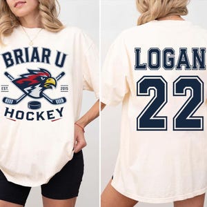 Puede incluir: Camiseta color crema con dise&ntilde;o de hockey. La parte delantera muestra "BRIAR U HOCKEY" con palos de hockey cruzados, un logotipo y el a&ntilde;o "2015". La parte trasera muestra "LOGAN 22" en letras may&uacute;sculas azul marino.