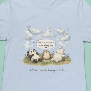May include: Light blue t-shirt featuring a whimsical illustration of a panda, hedgehog, and rabbit lounging on grass, with cloud-shaped animals above. Text reads "the sky puts on a show every day" and "cloud watching club."