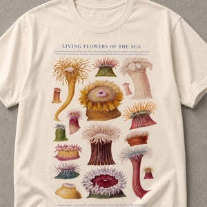 以下が含まれることがあります： LIVING FLOWERS OF THE SEAというタイトルのヴィンテージ風の科学イラストが描かれたクリーム色のTシャツ。黄色、ピンク、赤、緑の色合いの様々なカラフルなイソギンチャクが描かれており、小さな番号付きのラベルが付いています。