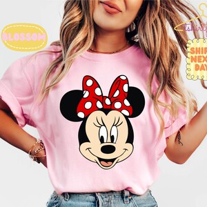 May include: A light pink t-shirt featuring a Minnie Mouse graphic. Minnie Mouse has a black head, ears, and a red bow with white polka dots. The shirt has rolled-up sleeves and the text "BLOSSOM" and "SHIPS NEXT DAY" are visible.