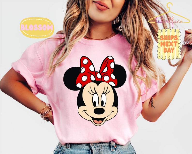 May include: A light pink t-shirt featuring a Minnie Mouse graphic. Minnie Mouse has a black head, ears, and a red bow with white polka dots. The shirt has rolled-up sleeves and the text "BLOSSOM" and "SHIPS NEXT DAY" are visible.