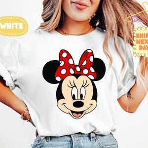 May include: White t-shirt featuring a cartoon illustration of Minnie Mouse's face. Minnie has black ears, a red bow with white polka dots, and a smiling expression. The word "WHITE" is in a yellow bubble.