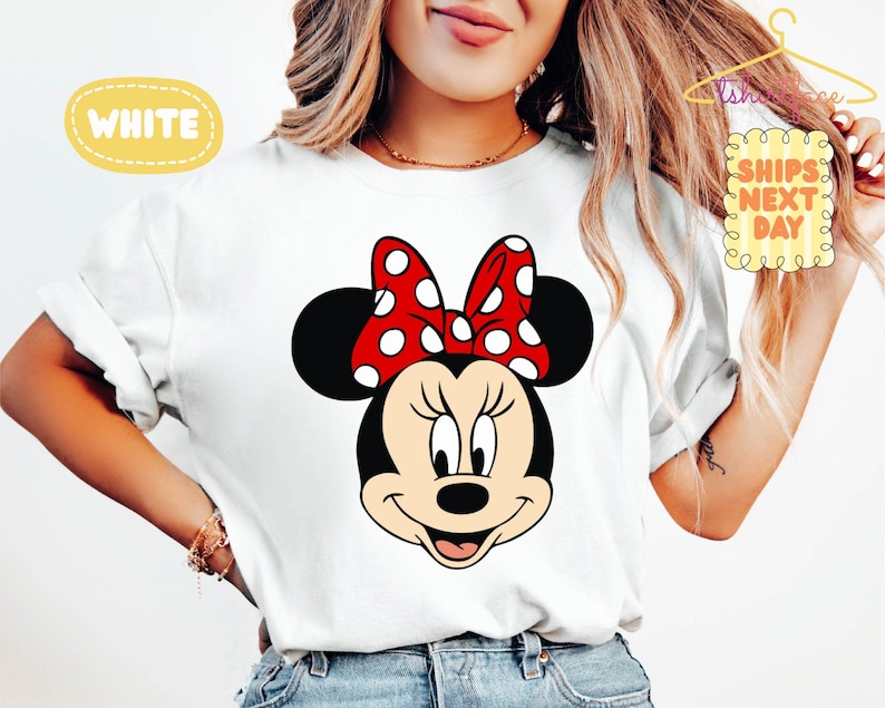 May include: White t-shirt featuring a cartoon illustration of Minnie Mouse's face. Minnie has black ears, a red bow with white polka dots, and a smiling expression. The word "WHITE" is in a yellow bubble.
