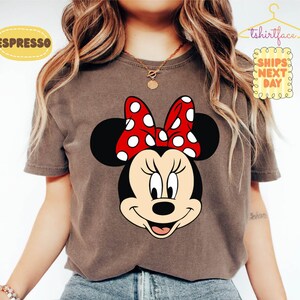 May include: Espresso-colored t-shirt featuring a large graphic of Minnie Mouse's face. Minnie has a red bow with white polka dots. The shirt has the text "tshirtface" and "SHIPS NEXT DAY" on it.