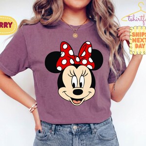 May include: Berry-colored t-shirt featuring a Minnie Mouse graphic. The graphic shows Minnie's face with a red bow with white polka dots. The shirt has short sleeves and is made of a soft material. The text "SHIPS NEXT DAY" is also visible.