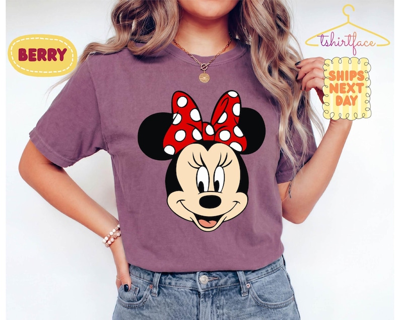 May include: Berry-colored t-shirt featuring a Minnie Mouse graphic. The graphic shows Minnie's face with a red bow with white polka dots. The shirt has short sleeves and is made of a soft material. The text "SHIPS NEXT DAY" is also visible.