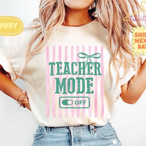 以下が含まれることがあります： アイボリー色のTシャツ。緑色のブロック体で「TEACHER MODE OFF」と書かれています。デザインにはピンクと白のストライプの背景と、ティール色のリボンが含まれています。カジュアルなスタイルです。