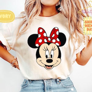 May include: Ivory-colored t-shirt featuring a large graphic of Minnie Mouse's face. Minnie's face is black with a red bow with white polka dots. The shirt also has the text "SHIPS NEXT DAY".