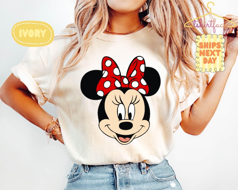 May include: Ivory-colored t-shirt featuring a large graphic of Minnie Mouse's face. Minnie's face is black with a red bow with white polka dots. The shirt also has the text "SHIPS NEXT DAY".