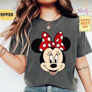 May include: Dark gray t-shirt featuring a Minnie Mouse graphic. Minnie Mouse is depicted with a black head, ears, and a red bow with white polka dots. The shirt has the text "Pepper" and "Ships Next Day".
