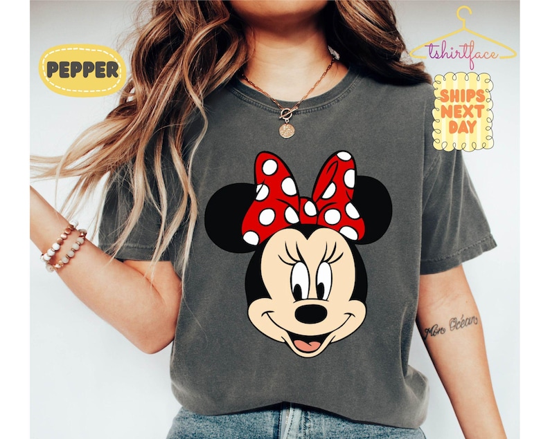 May include: Dark gray t-shirt featuring a Minnie Mouse graphic. Minnie Mouse is depicted with a black head, ears, and a red bow with white polka dots. The shirt has the text "Pepper" and "Ships Next Day".