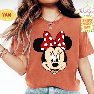 May include: A rust-colored t-shirt featuring a large graphic of Minnie Mouse's face with a red polka-dot bow. The shirt has short sleeves and a crew neck. The text "YAM" is in a yellow oval on the left side of the shirt. The text "tshirtface" is above the right shoulder.