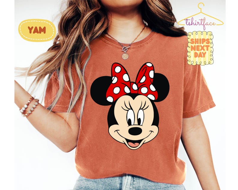 May include: A rust-colored t-shirt featuring a large graphic of Minnie Mouse's face with a red polka-dot bow. The shirt has short sleeves and a crew neck. The text "YAM" is in a yellow oval on the left side of the shirt. The text "tshirtface" is above the right shoulder.
