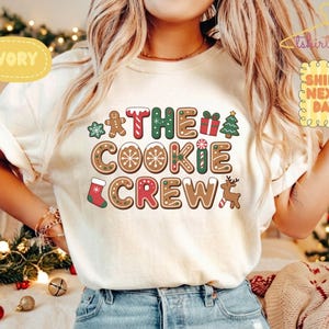 Może przedstawiać: Koszulka z krótkim rękawem w kolorze kości słoniowej z nadrukiem graficznym z napisem THE COOKIE CREW w stylu piernikowych liter. Wzór zawiera ilustracje piernikowych ludzików, choinki, prezentu, świątecznej skarpety oraz renifera.