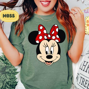May include: Moss green t-shirt featuring a Minnie Mouse graphic. The graphic shows Minnie's face with a red bow with white polka dots. The shirt has a crew neck and short sleeves. The text "MOSS" is in a yellow oval.