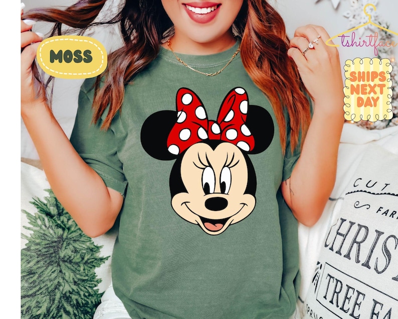 May include: Moss green t-shirt featuring a Minnie Mouse graphic. The graphic shows Minnie's face with a red bow with white polka dots. The shirt has a crew neck and short sleeves. The text "MOSS" is in a yellow oval.