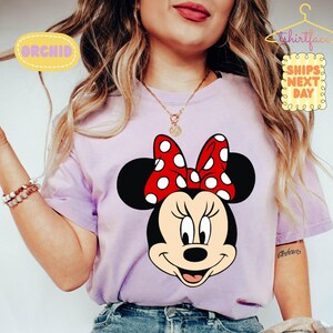 May include: A light purple t-shirt featuring a large graphic of Minnie Mouse. Minnie Mouse has a red bow with white polka dots. The shirt has the text "ORCHID" and "SHIPS NEXT DAY" on it.