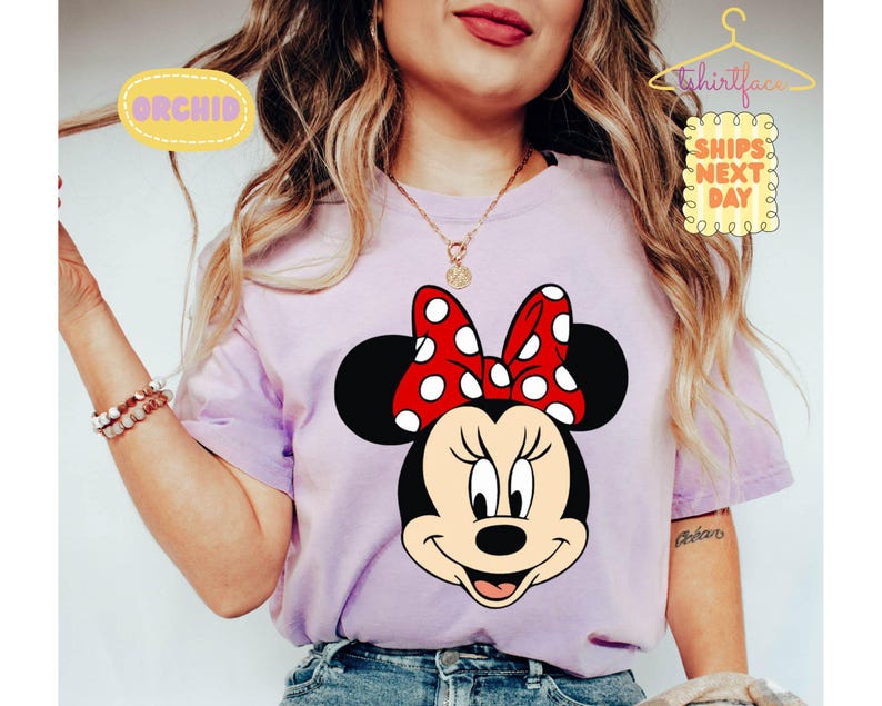 May include: A light purple t-shirt featuring a large graphic of Minnie Mouse. Minnie Mouse has a red bow with white polka dots. The shirt has the text "ORCHID" and "SHIPS NEXT DAY" on it.