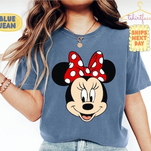 May include: Blue t-shirt featuring a Minnie Mouse graphic. The graphic shows Minnie's face with a red bow with white polka dots. The shirt has the text "BLUE JEAN" and "tshirtface SHIPS NEXT DAY".