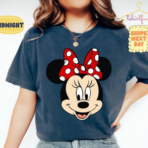 May include: A navy blue t-shirt featuring a large graphic of Minnie Mouse's face. Minnie Mouse has a black head, a red bow with white polka dots, and a smiling expression. The text "Midnight" is in a yellow oval.