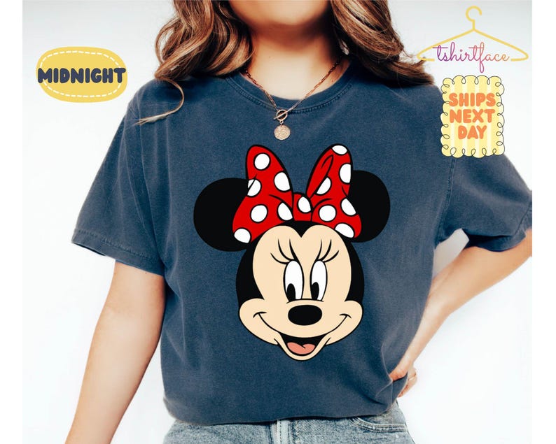May include: A navy blue t-shirt featuring a large graphic of Minnie Mouse's face. Minnie Mouse has a black head, a red bow with white polka dots, and a smiling expression. The text "Midnight" is in a yellow oval.