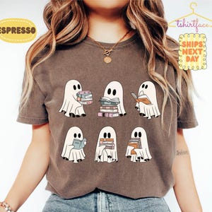 May include: A dark brown short-sleeved t-shirt featuring a graphic print of six white ghosts, each holding or reading stacks of colourful books. The ghosts are arranged in two rows, with each figure depicted in a simple, cartoon-like style.